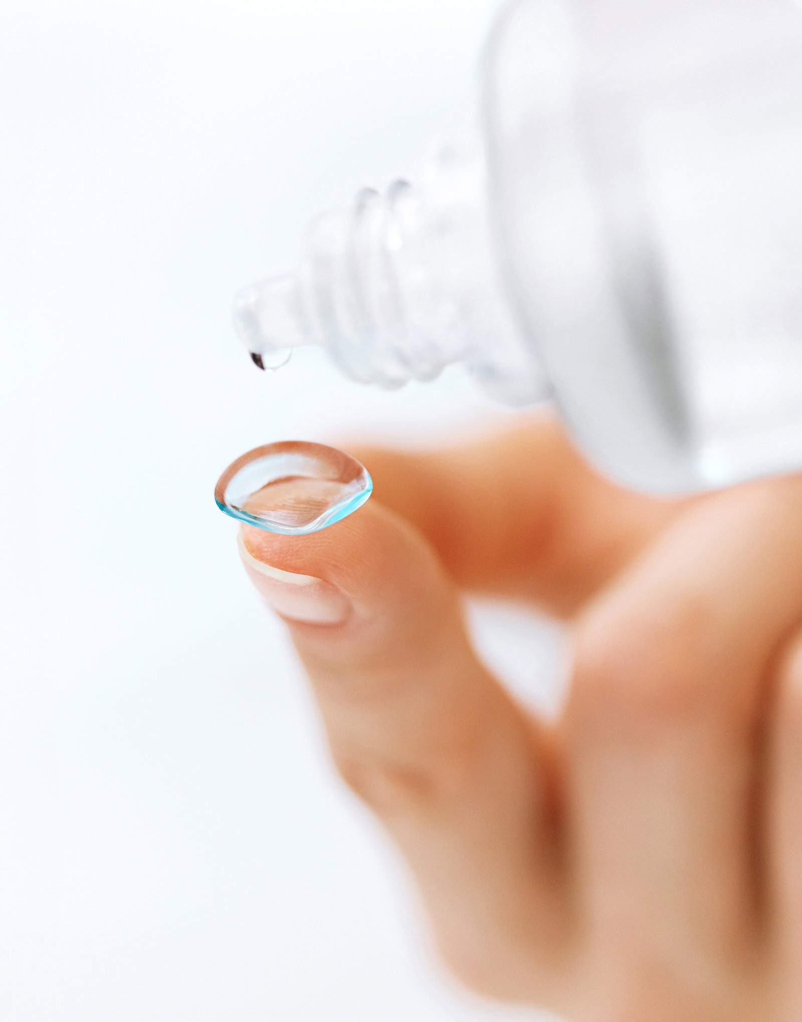 Das Bild zeigt Finger mit einer Linse und eine Flasche Linsenpflegemittel.