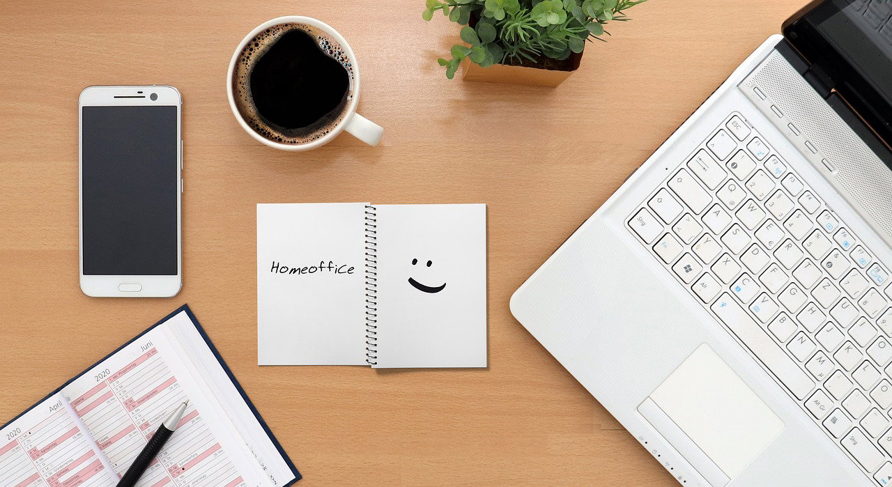 Zu sehen ist ein Schreibtisch mit Laptop, IPhone, Kalender, Block und Tasse.