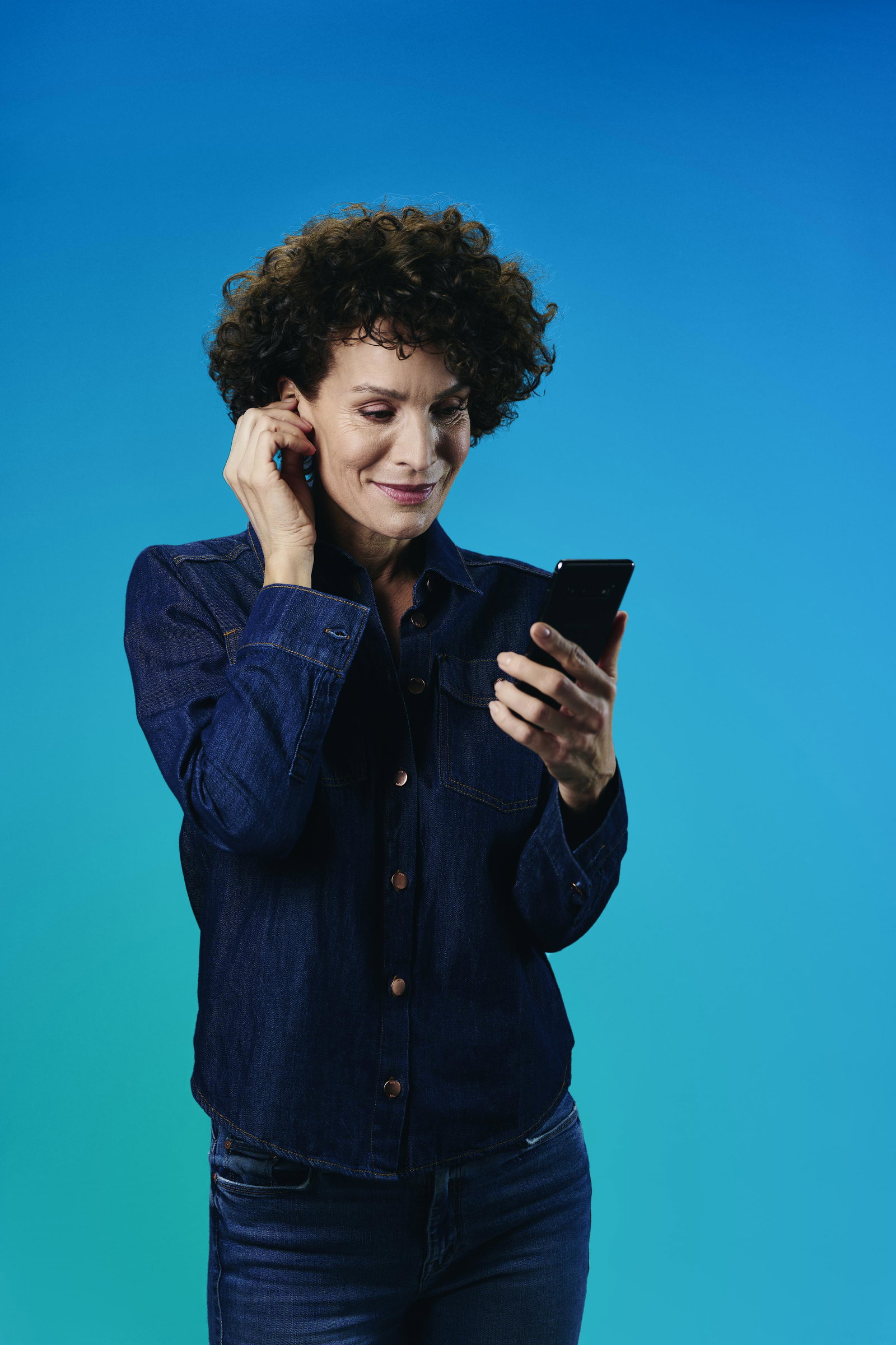 Das Bild zeigt eine Frau vor, die eine Hand am Ohr hat und auf ein Handy schaut.