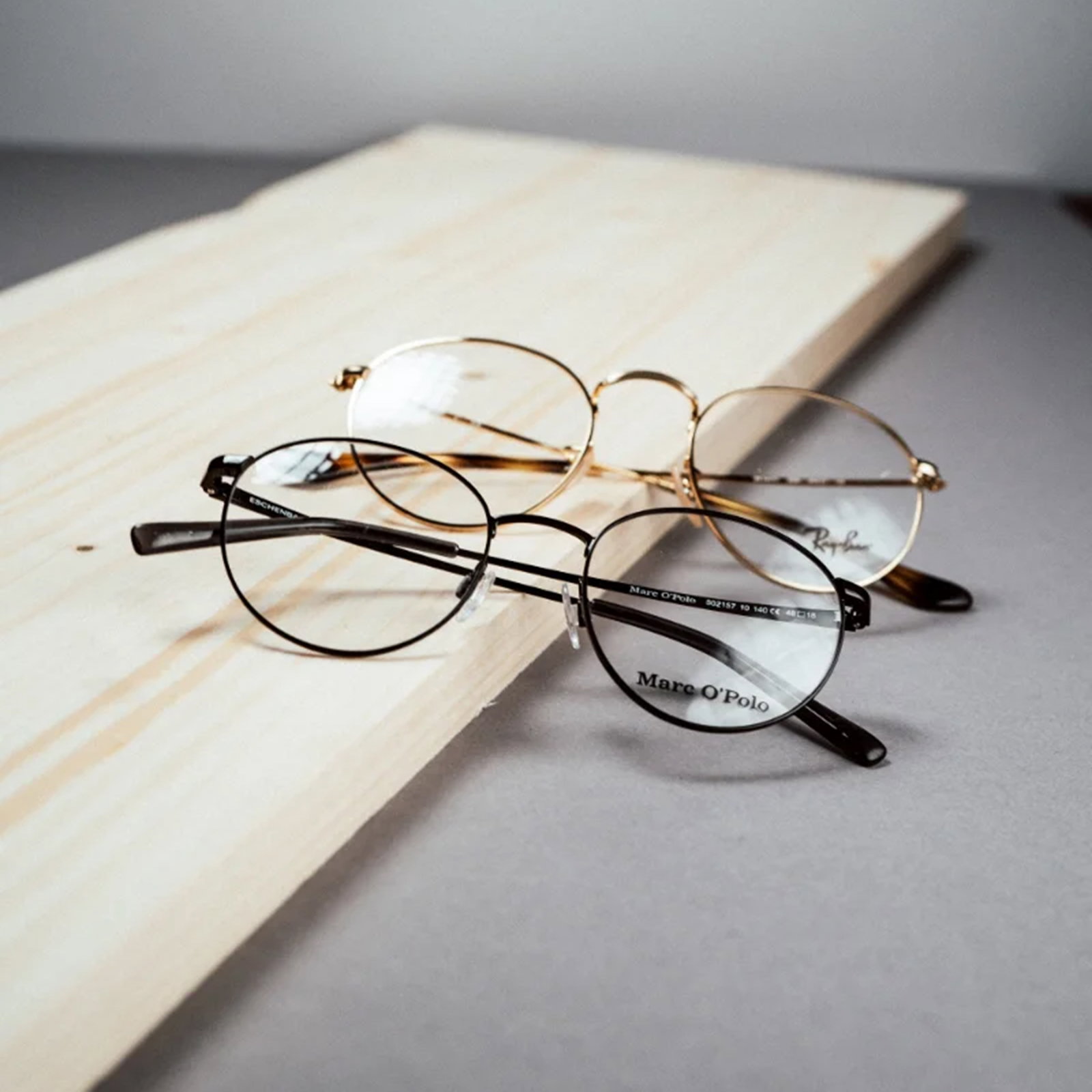Das Bild zeigt eine schwarze und  eine goldene Metallbrille, die hintereinander auf einem Holzbrett liegen.